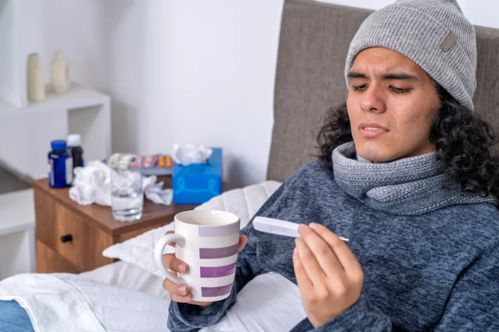 Person resting in bed with a thermometer and warm drink, experiencing a returning fever after missing a dose of Amoxicillin